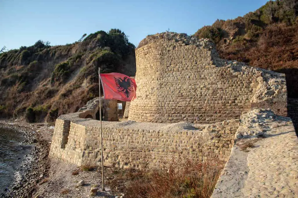 planeando un viaje a Albania, paredes de piedra blanca del castillo Rodoni con bandera albanesa roja y negra ondeando en el viento con acantilados costeros detrás