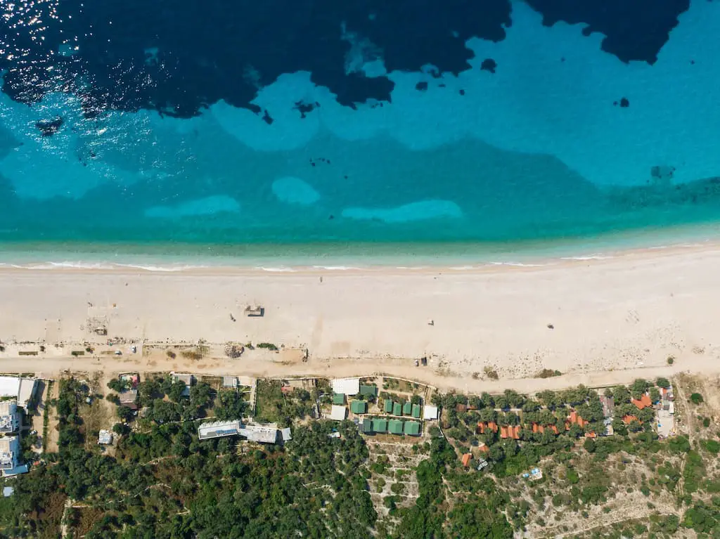 Disfrute del último itinerario de una semana en Albania, vista aérea de la playa de arena blanca y aguas cristalinas de color azul turquesa con varias áreas de edificios y árboles cerca. qué ver en albania en 7 días