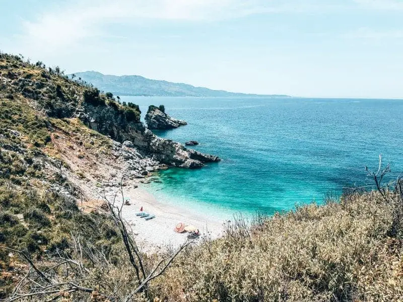 Encuentre las mejores ciudades para visitar en Albania para viajes a la playa, área de césped que conduce a una pequeña área de playa con tumbonas y agua clara con colinas en la distancia