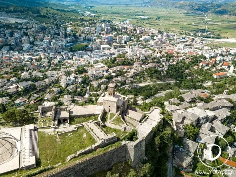 10 ciudades increíbles en Albania para explorar, toma aérea de fortificaciones históricas con torre de reloj de piedra con vistas a la ciudad con muchos edificios modernos junto a un río con campos verdes en la distancia