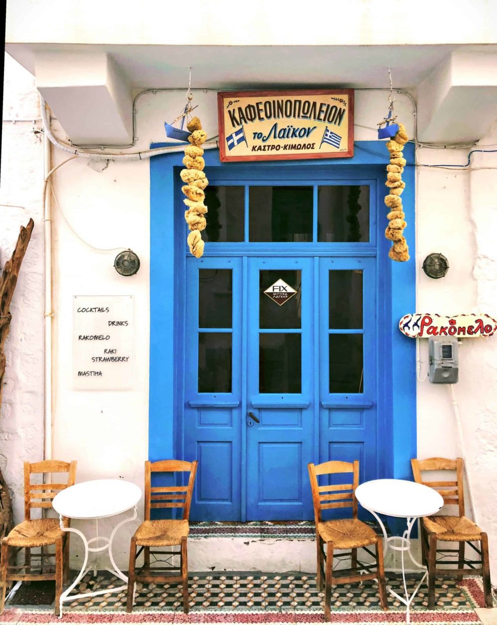 10 platos griegos tradicionales para probar en Grecia, frente a un café griego con una gran puerta de madera azul detrás de pequeñas mesas con sillas de mimbre en la calle