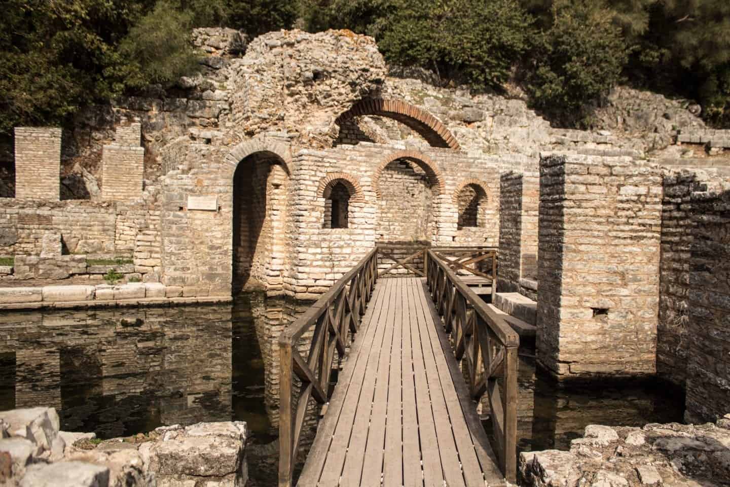 Castillos en Albania para disfrutar, puente de madera que atraviesa el agua a través de ruinas de paredes de piedra blanca y arcos con vegetación detrás