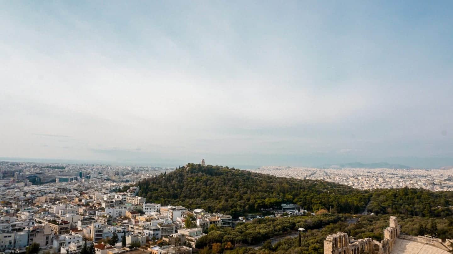 Encuentre los lugares más bellos de Grecia, vista del paisaje urbano de Atenas con una gran área construida de edificios que rodean la colina cubierta densamente de árboles verdes