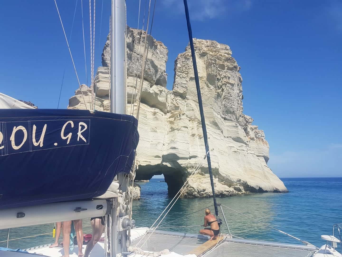 Descubre los mejores lugares para ver en Grecia, personas sentadas en un bote navegando frente a una gran formación rocosa blanca sentada en aguas tranquilas y azules
