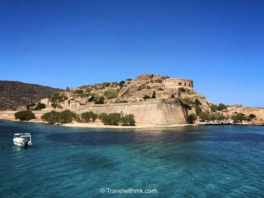 ¿Has estado en estos lugares únicos para visitar en Grecia? Antiguas fortificaciones de piedra situadas en una playa de arena junto a aguas cristalinas con un barco en primer plano bajo un cielo azul celeste.