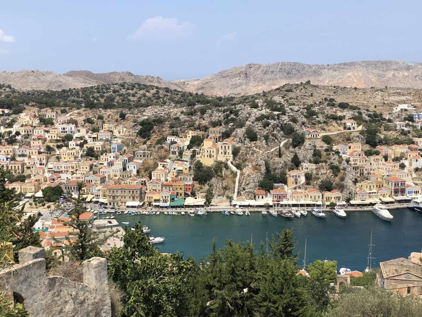 Venga y vea el mejor lugar para visitar en Grecia, vista del puerto con edificios que bordean el agua y colinas cubiertas de hierba seca que conducen a la distancia