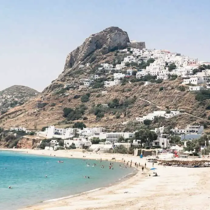 Explore las mejores ciudades para visitar en Grecia, colina alta con edificios que conducen a la cima entre praderas marrones y árboles verdes con playa de arena blanca y agua azul en primer plano