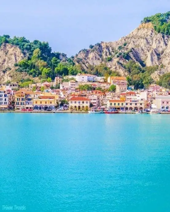 Encuentre la mejor ciudad de Grecia para visitar para los amantes de la comida, una foto de la ciudad con edificios coloridos ubicados entre grandes colinas rocosas sobre aguas azules vibrantes