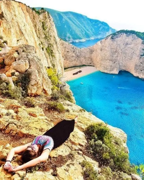 Encuentre sus destinos populares favoritos en Grecia, persona acostada sobre el borde del acantilado con vistas a un lago turquesa claro con playa de arena
