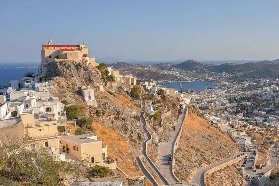 Encuentre las mejores ciudades de Grecia para visitar este verano, una colina alta con un gran edificio en lo alto que mira hacia la ciudad debajo con colinas y el mar en la distancia