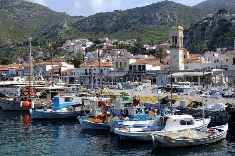 Disfruta de los mejores lugares para ir en Grecia, puerto con varios barcos amarrados junto a un pequeño pueblo frente a verdes colinas rocosas