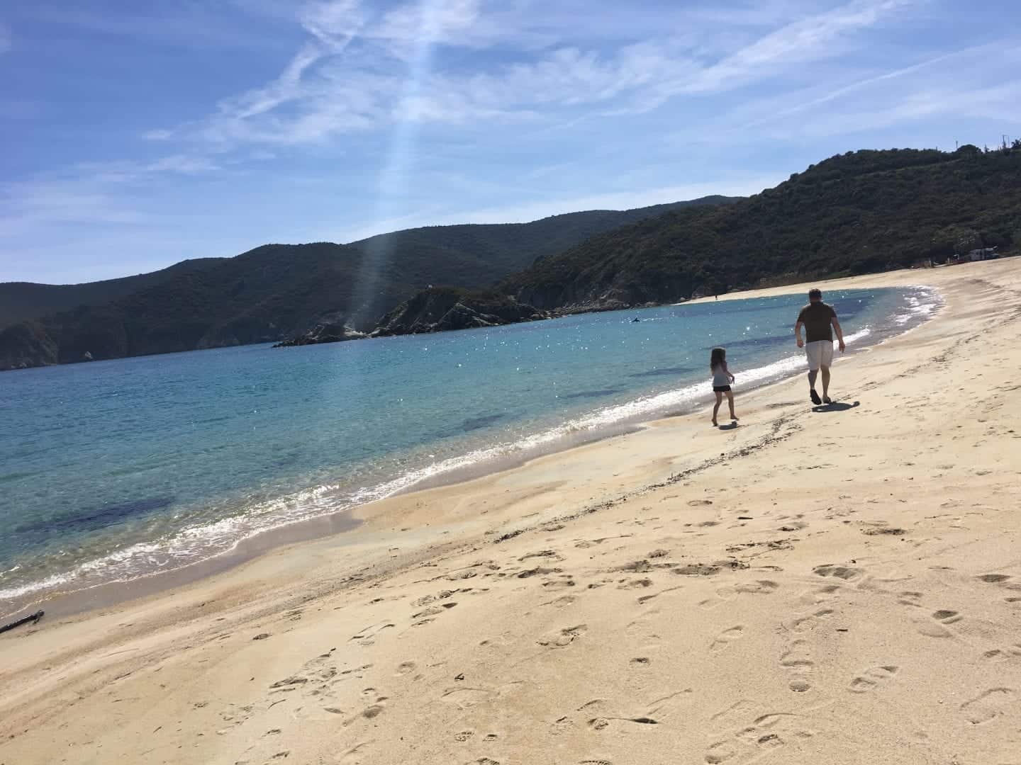 No te pierdas ninguna de estas increíbles cosas para ver en Grecia, dos personas caminando por la playa de arena mientras las olas golpean suavemente la orilla bajo un sol brillante con colinas en la distancia.