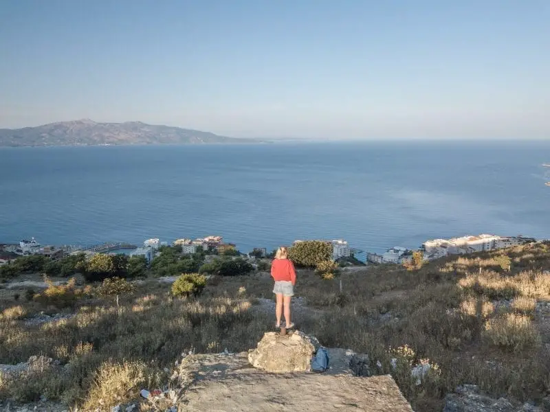 Los mejores consejos de viaje de Albania, una persona con un puente rojo sobre una roca mirando hacia una gran masa de agua azul con una montaña a lo lejos