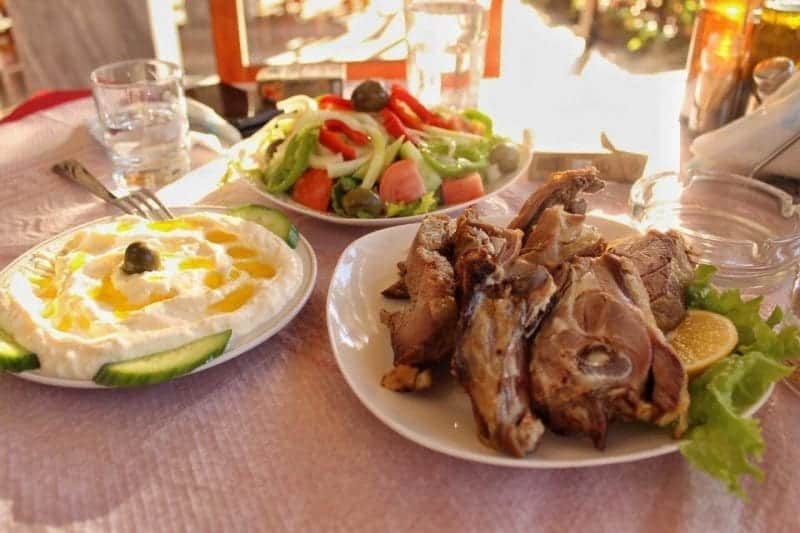Consejos para viajar en Albania, mesa cubierta con varios deliciosos platos tradicionales albaneses que incluyen ensalada de verduras de pimientos y tomates y carne en el hueso con guarnición de naranja