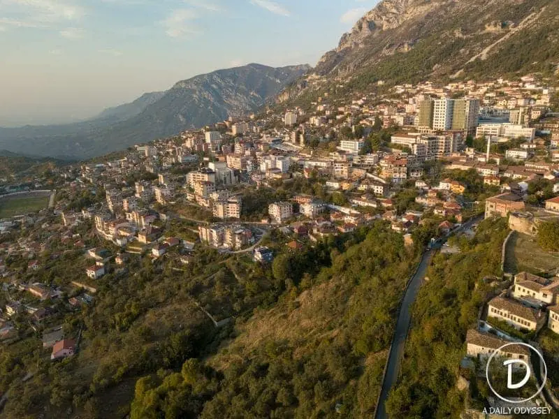 Explore la hermosa ciudad de Kruja, toma aérea de la ciudad con edificios que bordean la ladera inclinada entre los árboles y las áreas verdes que conducen alrededor de la curva de la colina hacia las montañas brumosas en la distancia