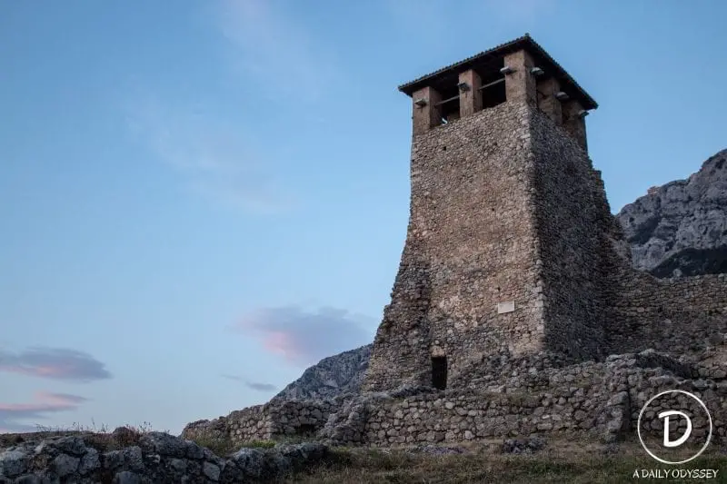 Viaje al castillo de Kruja, alta torre de piedra con restos del muro de piedra circundante y montañas detrás bajo un cielo crepuscular