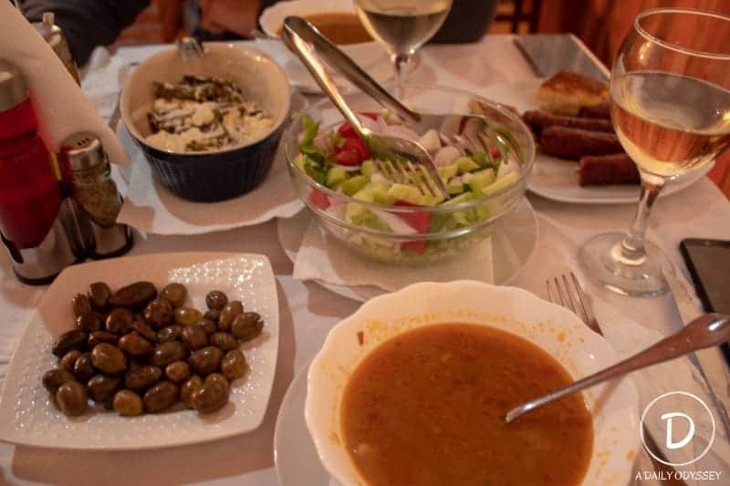 Disfrute de hoteles económicos en Kruje con un presupuesto ajustado, plato de ensalada con pinzas plateadas junto a plato de sopa especiada y plato de aceitunas marinadas y plato de salchichas con una copa de vino blanco y sal y pimienta sobre un mantel blanco