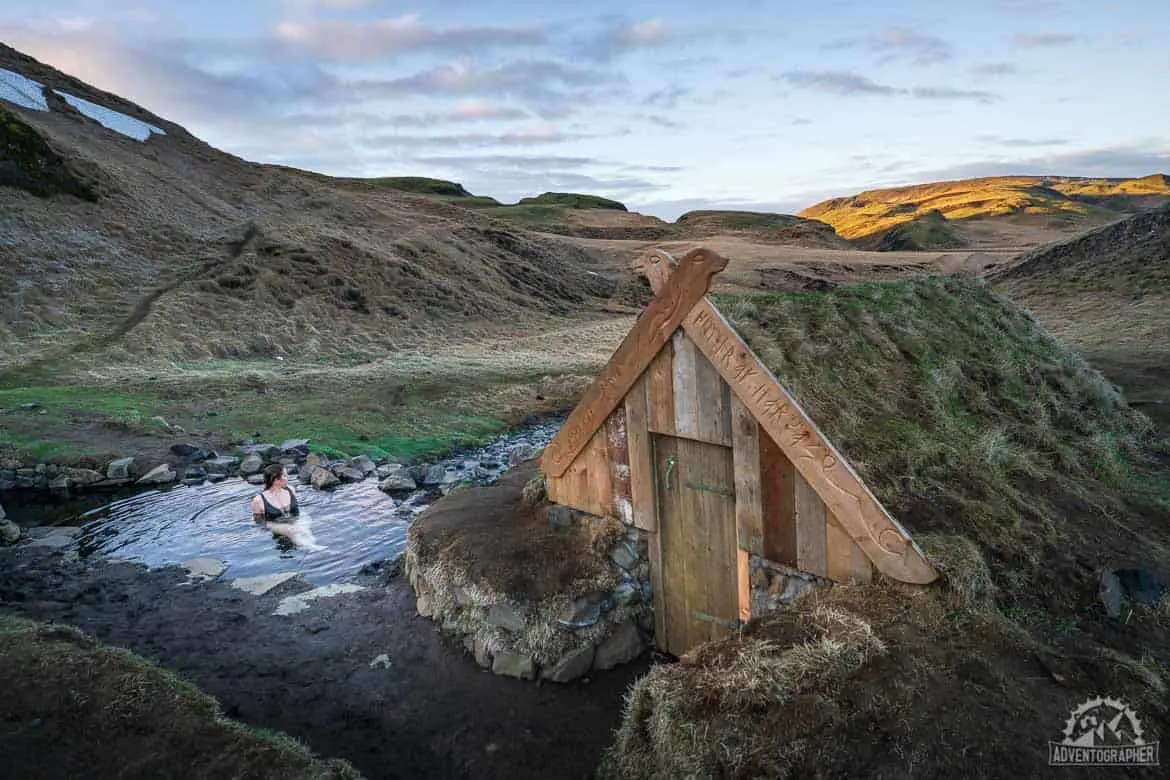 Qué hacer en Vik Islandia, cabaña de madera con techo de pasto orgánico con un estanque rocoso de agua cerca en el que está sentada una persona en traje de baño con colinas cubiertas de hierba al fondo