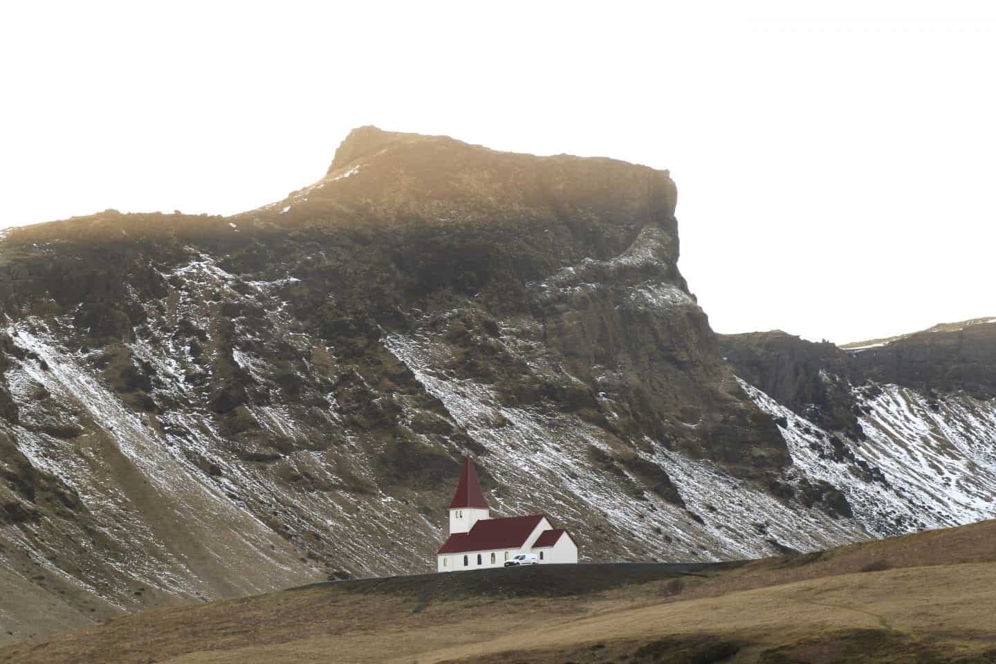 Debe hacer actividades en Vik Islandia, iglesia blanca con techo de tejas rojas sentada en la cima de una colina frente a un gran afloramiento rocoso con algo de nieve a los lados bajo un cielo soleado y brillante