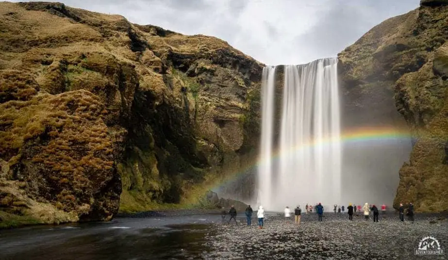 Qué hacer en Vik Islandia, hermosa cascada con varios arroyos de agua que caen por la ladera rocosa cubierta de musgo con un arco iris que se ve claramente en la neblina mientras varias personas miran desde el lecho del canal
