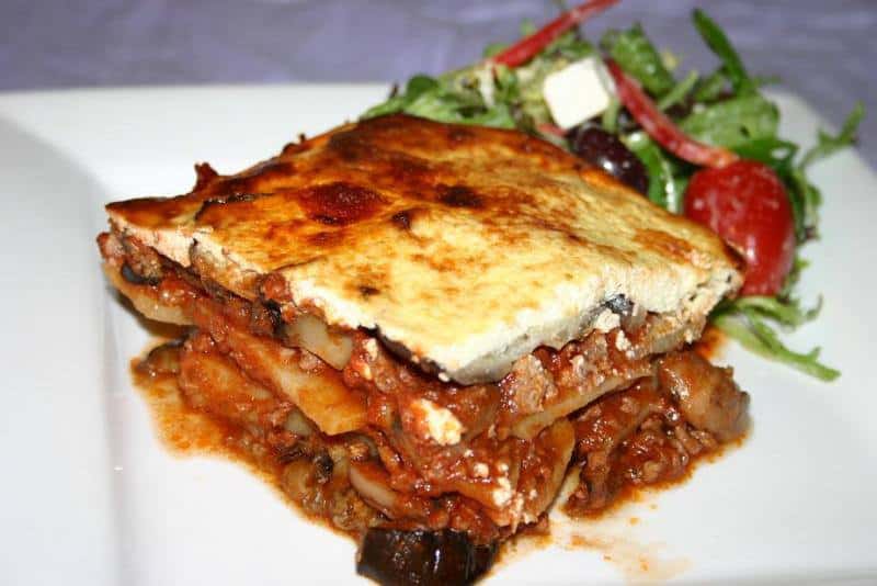 Experimente los platos tradicionales en Grecia, una rebanada de musaka con salsa y queso junto a la ensalada en un plato blanco