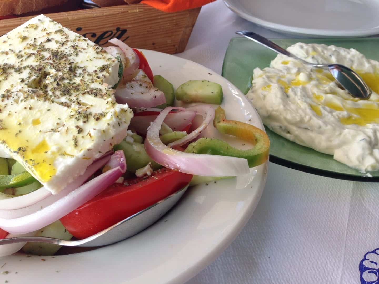 Disfrute de platos griegos fáciles, ensalada griega que incluye pimientos verdes, cebollas, tomates y queso feta en un plato blanco junto a un tazón de salsa cremosa