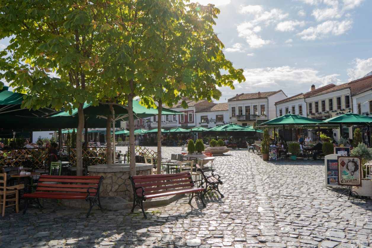 Qué hacer en Korca, calle adoquinada con bancos que rodean un pozo bajo árboles verdes con asientos cubiertos al aire libre para restaurantes que bordean la calle