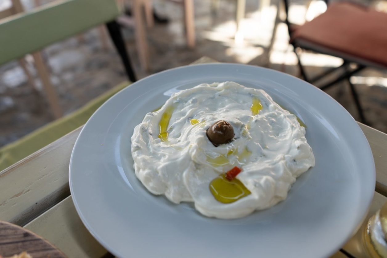 Qué comer en Korca Albania, salsa blanca cremosa adornada con aceitunas y aceite en un plato azul pálido sobre una mesa de madera verde con sillas