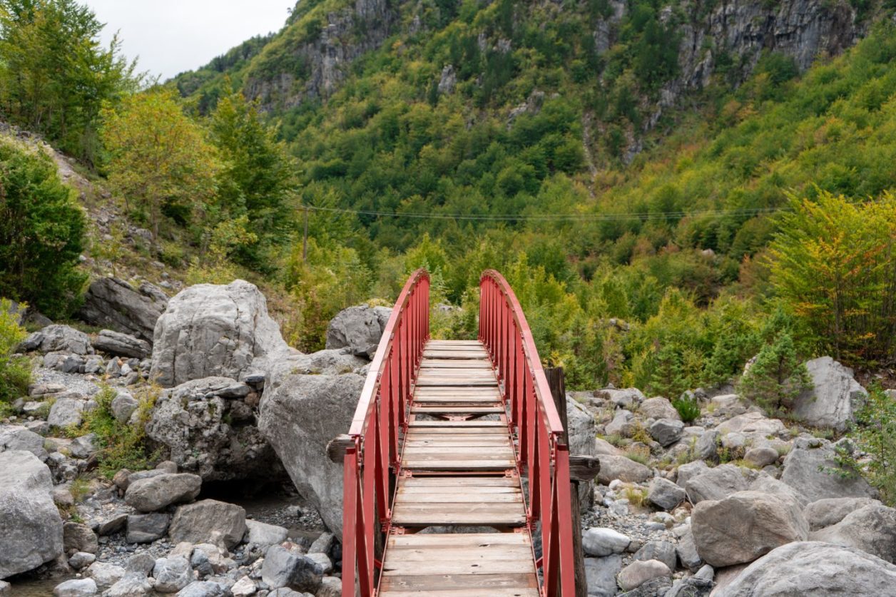 Explore la carretera de Theth, un puente de madera con barandillas rojas que cruza un río lleno de piedras con laderas rocosas verdes detrás