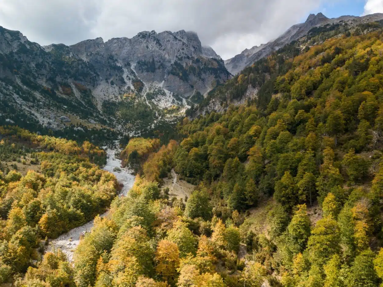 Explore el parque nacional Theth, toma aérea del río que serpentea a través de laderas cubiertas de árboles hacia montañas cubiertas de nubes en la distancia