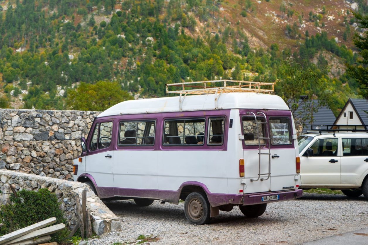 Descubra cómo llegar a Theth Albania, un minibús blanco con reflejos morados estacionado junto a un vehículo blanco de cuatro por cuatro frente a un muro de piedra con una colina cubierta de árboles en la distancia