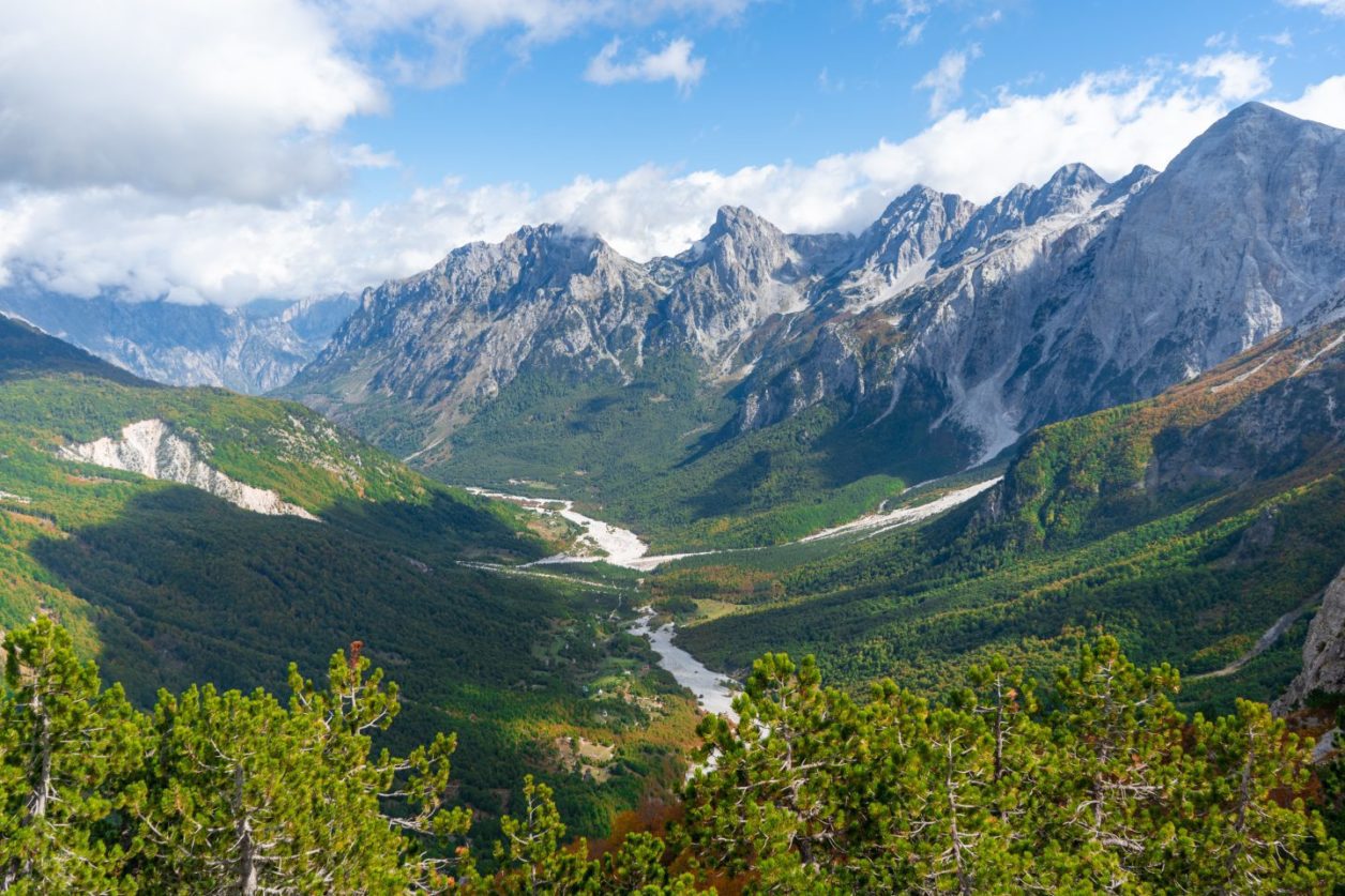 Visite la hermosa Theth Albania, vea el valle con laderas verdes y onduladas rodeadas de montañas rocosas cubiertas de nieve bajo un cielo azul con nubes