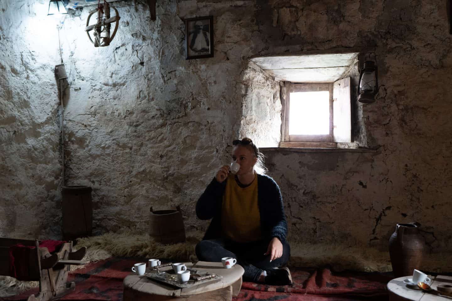 Cosas que hacer en Theth, Albania, tomando té dentro de una vivienda albanesa junto a una mesa baja con pequeñas tazas de té encima de una alfombra roja con ollas de almacenamiento cerca