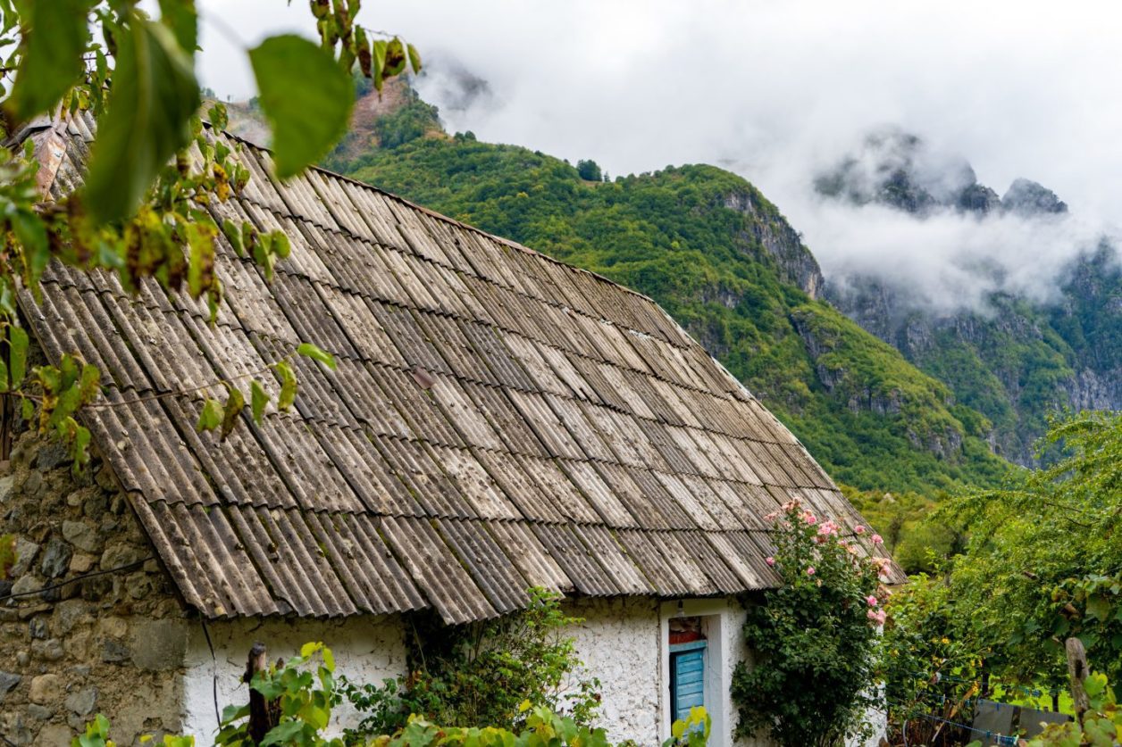 Cosas que hacer en Theth, Albania, casa blanca con techo de tejas grises frente a laderas cubiertas de follaje con picos oscurecidos por las nubes