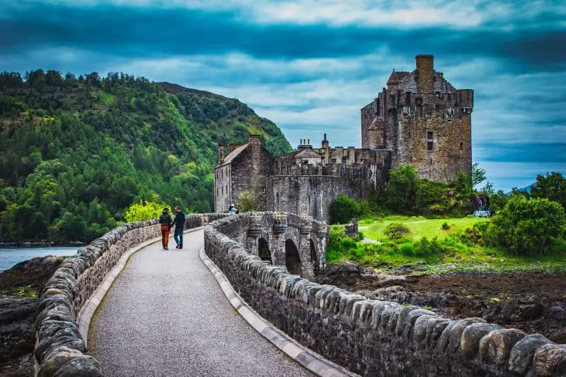 Vea estos mejores castillos en Europa, camino con paredes bajas de piedra que cruza un pequeño puente hacia un castillo de piedra gris con un campo verde y una colina boscosa detrás bajo un cielo nublado y cambiante