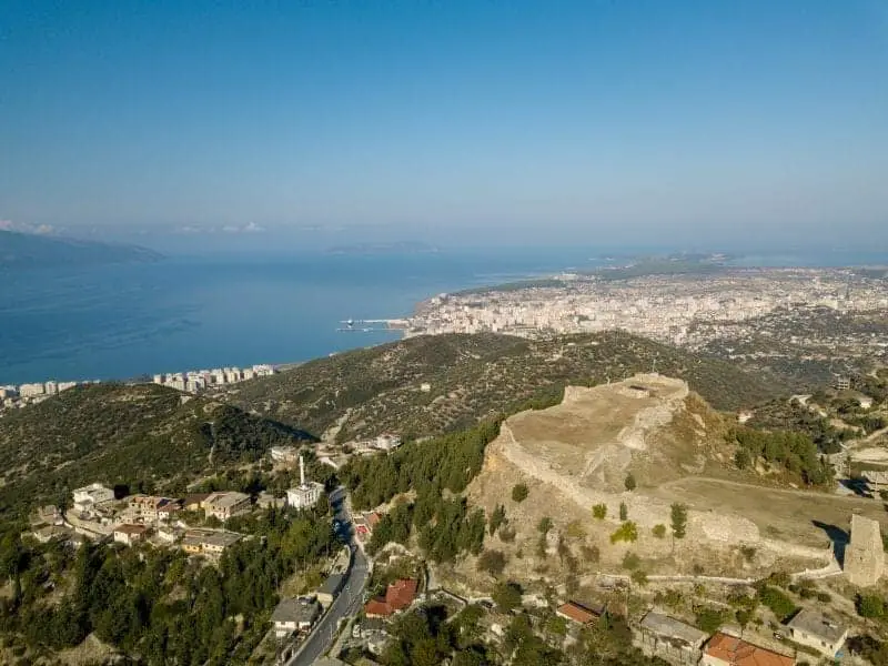 Explore hermosas ciudades albanesas, vista aérea de una gran zona de colinas con rocas y árboles que conducen hacia el paseo marítimo bajo un cielo azul
