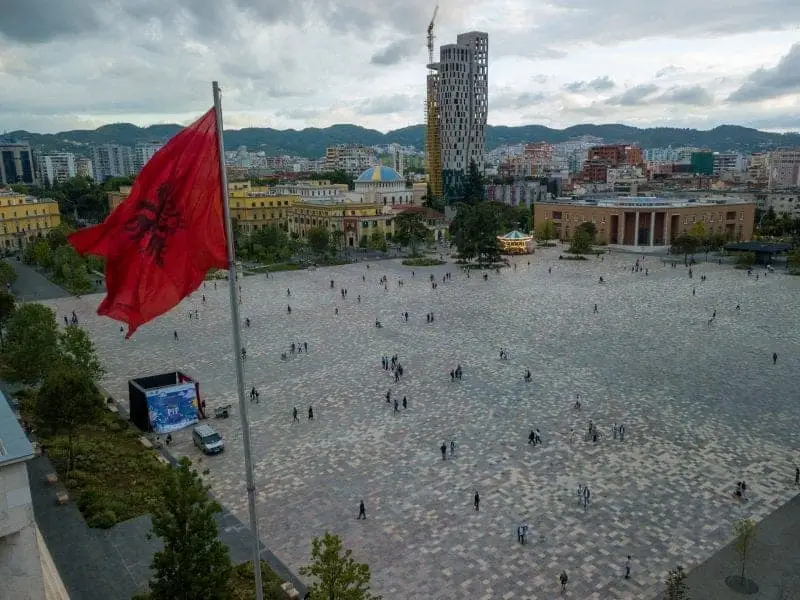 Descubra su nueva ciudad favorita de Albania, vista de la plaza pública principal de Tirana con gente esparcida como hormigas y la bandera nacional de Albania en un lugar destacado en primer plano