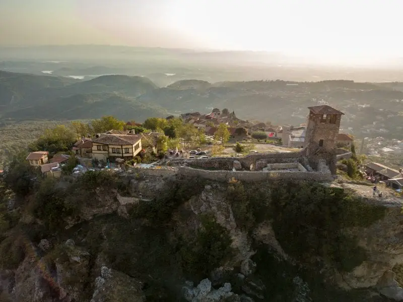 Visite las ciudades más grandes de Albania, un antiguo edificio de piedra con una torre y murallas fortificadas en lo alto de una gran colina rocosa con edificios más modernos cerca con colinas y valles en la distancia bajo el sol poniente