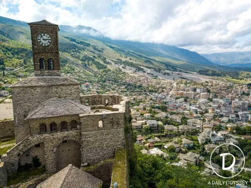 Las mejores ciudades de Albania para visitar este verano, la antigua torre del reloj de piedra con altos muros de piedra que miran desde lo alto de una colina hacia la ciudad moderna debajo con exuberantes colinas verdes y montañas en la distancia bajo un cielo nublado