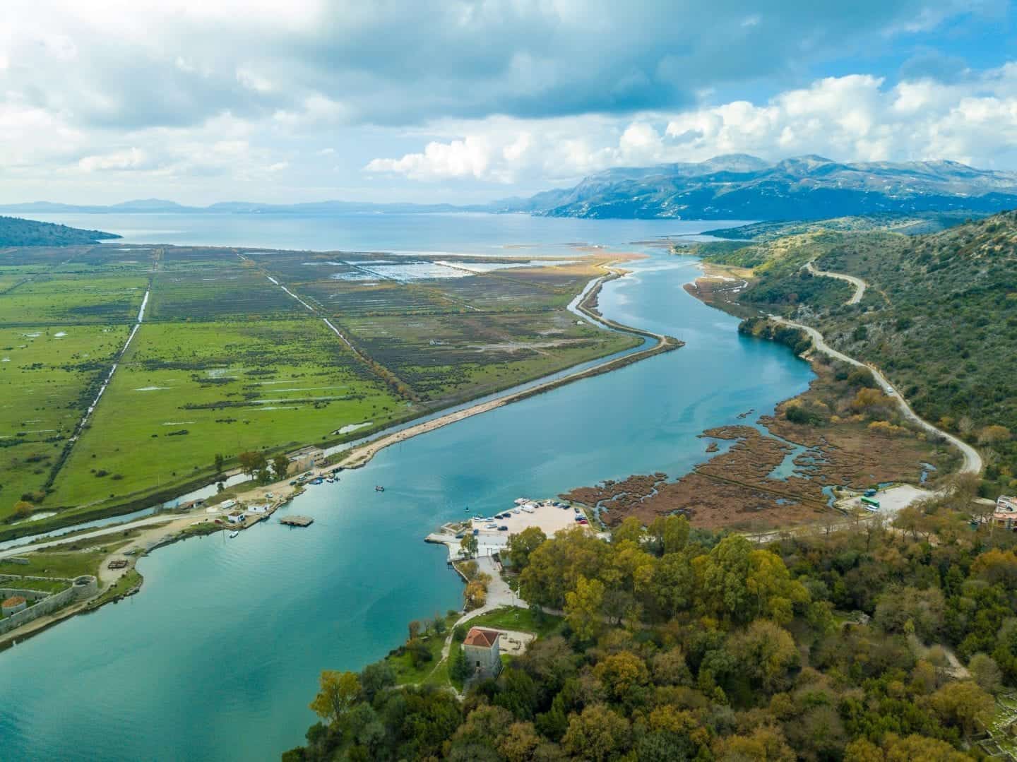 Descubra Butrint, Albania: uno de los mejores sitios de la UNESCO de Albania, vea Butrint, incluidas las áreas de playa y campo con un río que fluye hacia aguas abiertas con montañas en la distancia bajo un cielo nublado