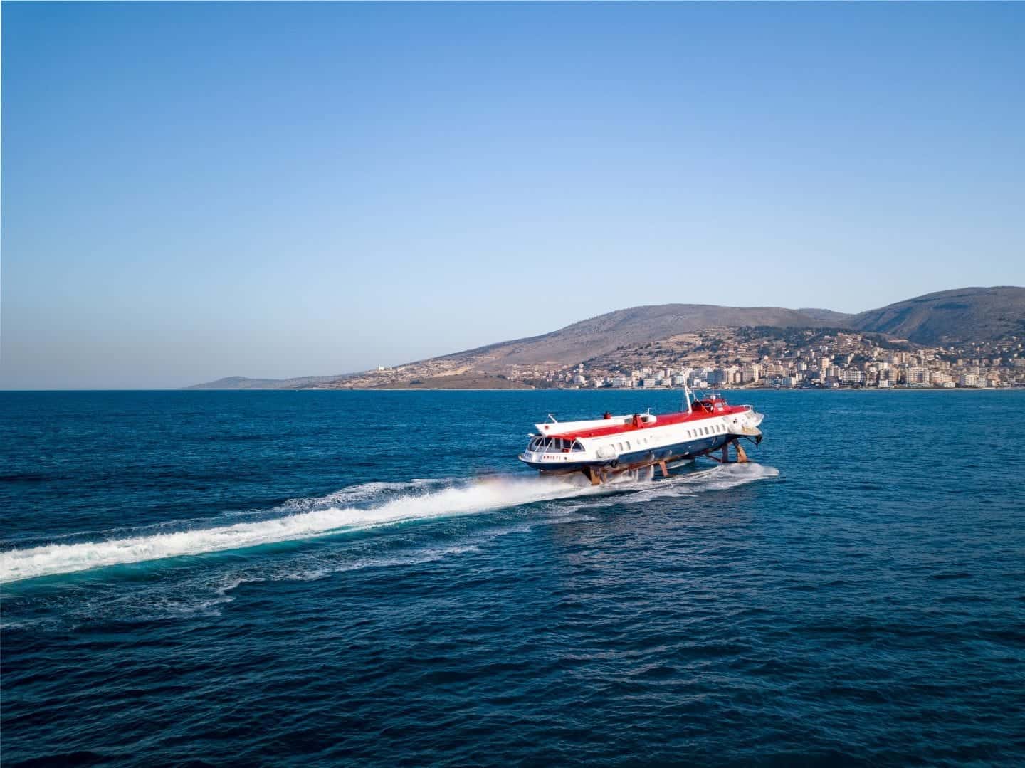 Una guía para hacer Albania en una semana, barco de hidroplano rojo, blanco y azul que navega a través del agua de mar azul profundo hacia una gran zona urbanizada con colinas detrás