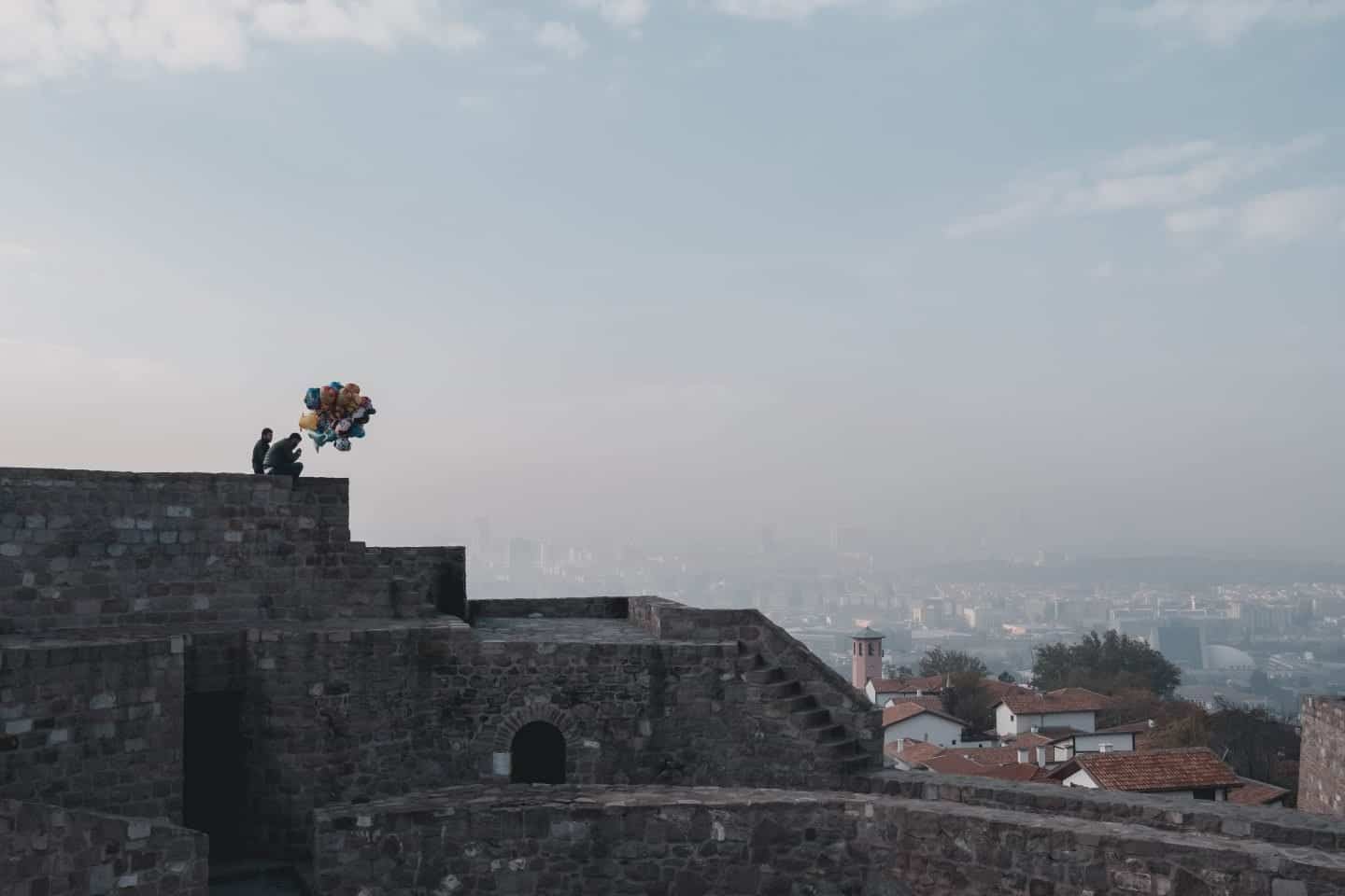 Impresionantes lugares para ver en Turquía, dos personas sosteniendo un montón de globos sentados sobre un muro de piedra frente a un telón de fondo de la ciudad de Ankara