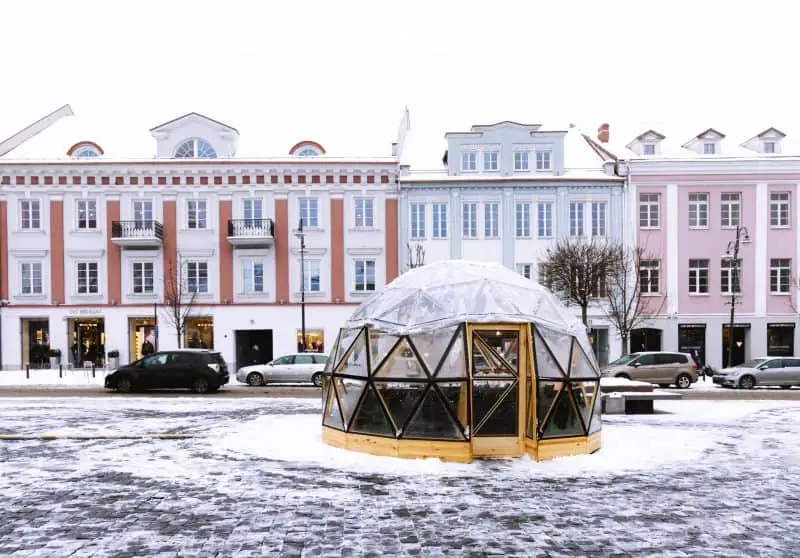 Encuentre los mejores lugares europeos más baratos para visitar, una plaza pública con una cúpula de cristal en el centro y una fila de autos estacionados afuera de unos edificios de tres pisos, todos cubiertos con una fina capa de blanco nieve