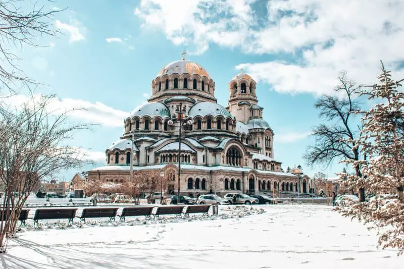 Explore los destinos europeos menos costosos, la estructura alta y compleja de la iglesia con muchas cúpulas y ventanas arqueadas con una línea de bancos de parque en primer plano y una capa de nieve sobre el paisaje
