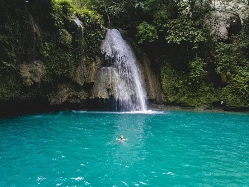 Playas subestimadas en Filipinas, persona nadando en una laguna turquesa junto a una cascada que baja por la roca junto al follaje verde