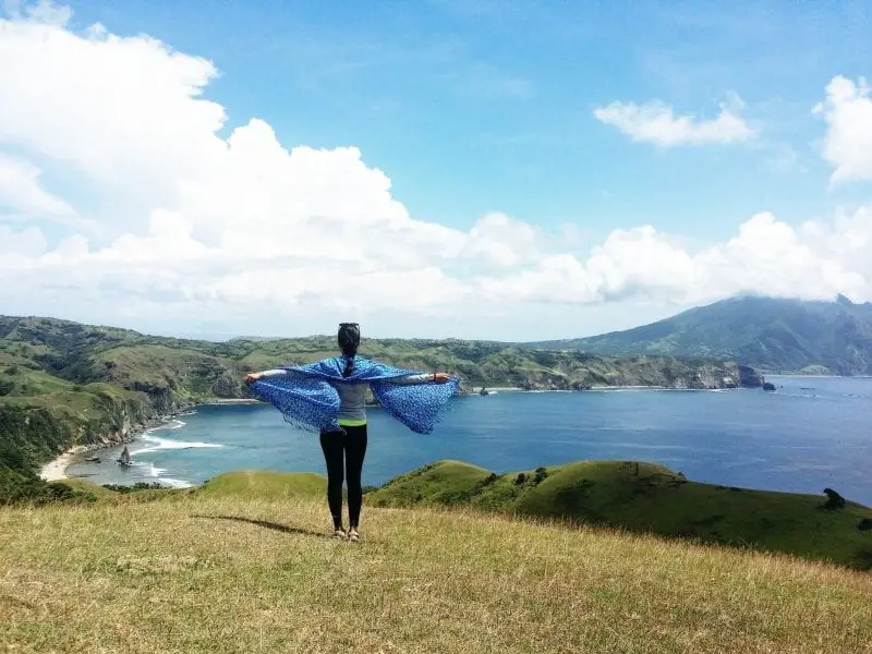 Bonita isla en Filipinas, mujer estirando los brazos de par en par hacia la entrada con playa y verdes colinas con montañas oscurecidas por nubes en la distancia