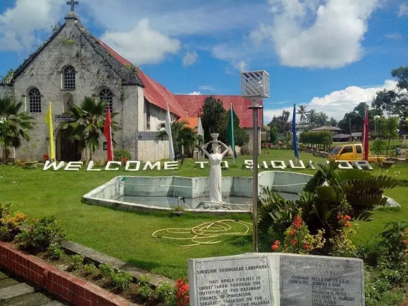 Las mejores islas de Filipinas, antiguo edificio de piedra con techo de tejas rojas con jardín que contiene césped y fuente con una estatua y letras deletreando 