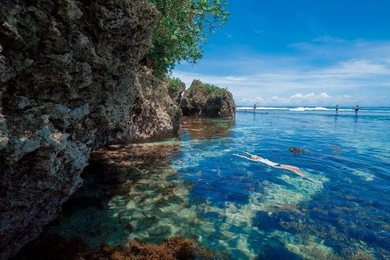 Las islas más hermosas de Filipinas, una persona nadando en aguas claras junto a paredes rocosas cubiertas de follaje y olas rompiendo en la distancia