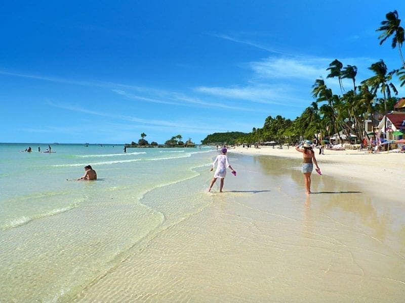Hermosas islas en Filipinas, gente caminando por aguas poco profundas en la playa sosteniendo sus zapatos con palmeras y cabañas de playa a un lado
