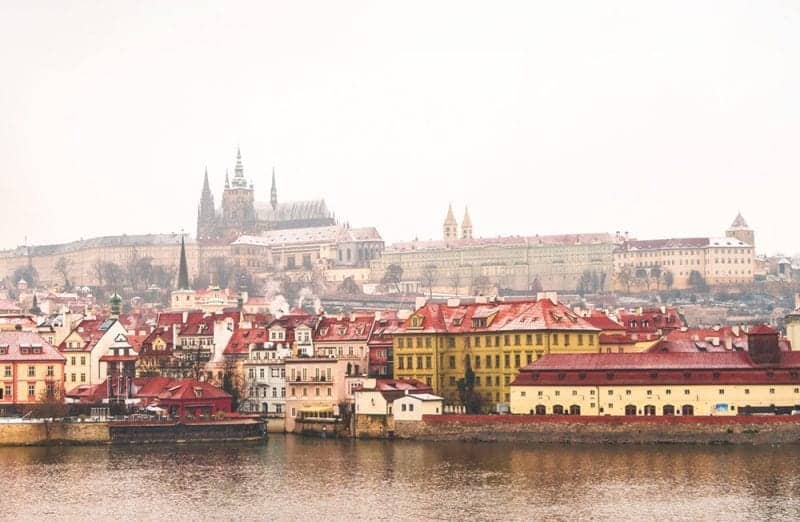Descubra las mejores ciudades europeas para visitar en enero, vista de Praga desde el río con grandes edificios europeos con torres puntiagudas y una iglesia en la cima de una colina con edificios de techos rojos agrupados a lo largo de la orilla del río en un día gris y nublado